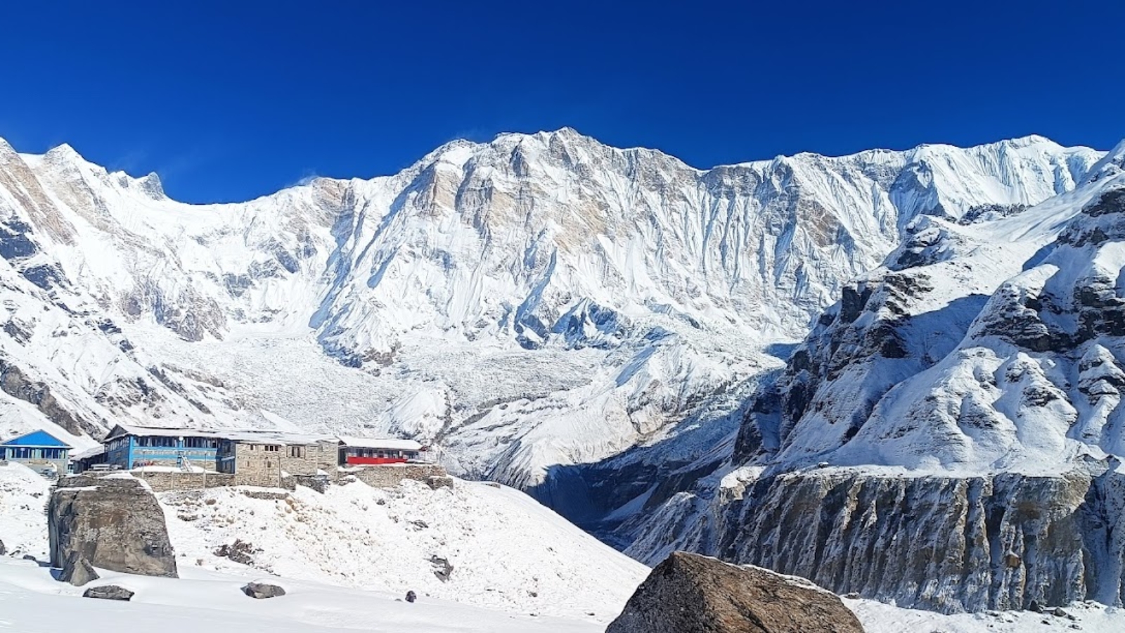 Snow-covered Annapurna Sanctuary with mountain lodges in the foreground, under a clear blue sky