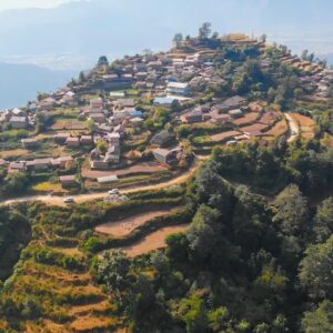 An aerial view of a rural village in Pokhara, Nepal, situated on a terraced hillside with traditional houses and lush greenery, overlooking distant valleys and mountains.