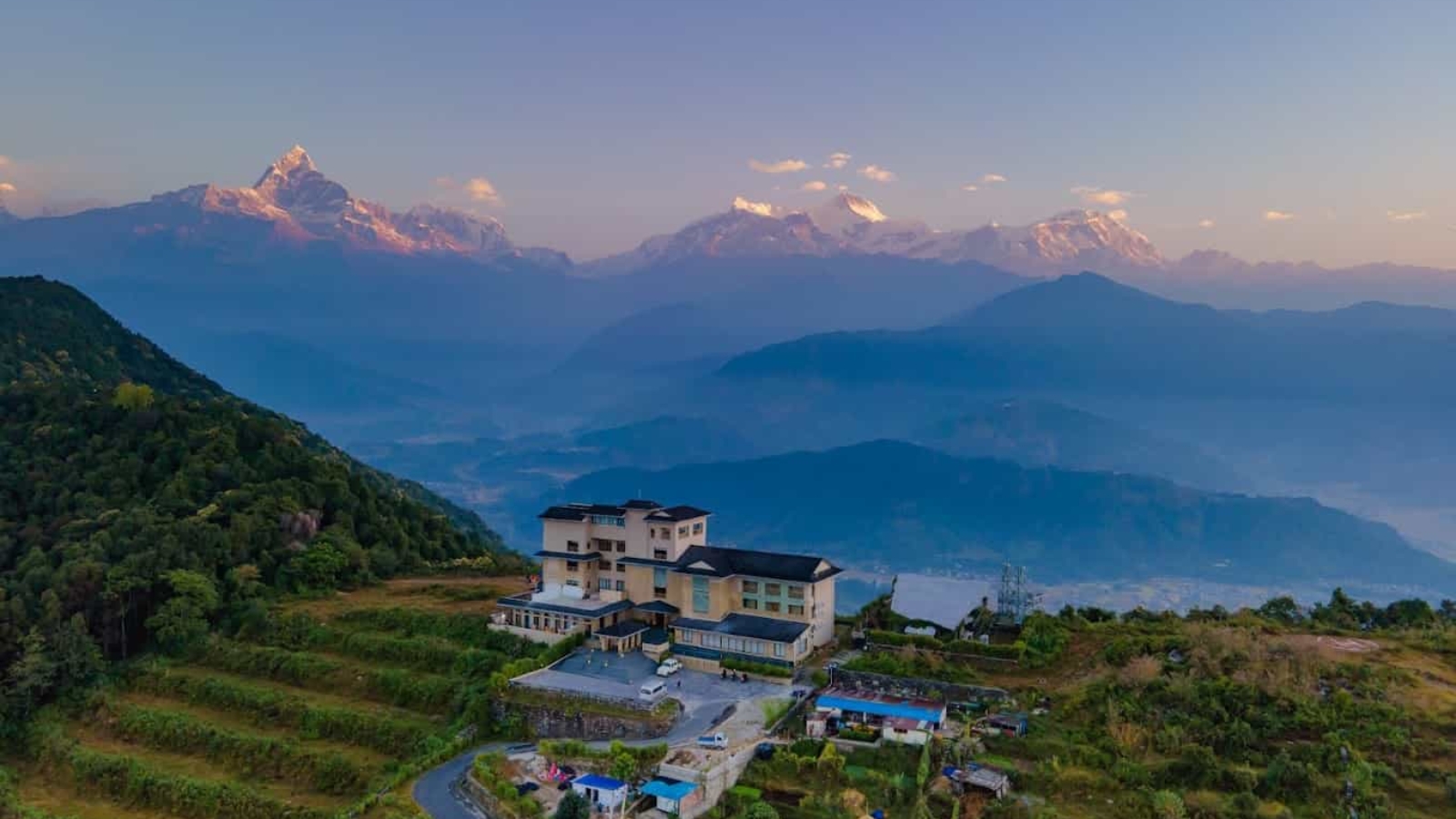 Panoramic view of a mountain resort surrounded by lush greenery with majestic Himalayan peaks in the background at sunrise.