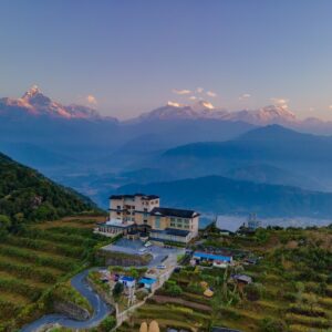 Panoramic view of a mountain resort surrounded by lush greenery with majestic Himalayan peaks in the background at sunrise.