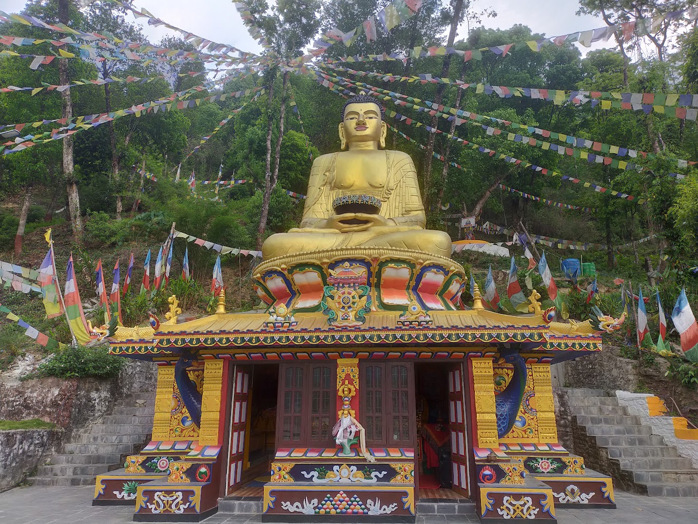 Golden Buddha statue at a vibrant monastery in Pokhara, Nepal, surrounded by colorful prayer flags and lush green trees, representing a key site on the Pokhara Buddhist Circuit.