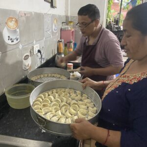 Two individuals preparing traditional Nepali momos in a kitchen, arranging them in large steam trays.