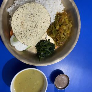 Traditional Nepali meal served on a brass plate with rice, curry, vegetables, papadum, and soup on a blue table.